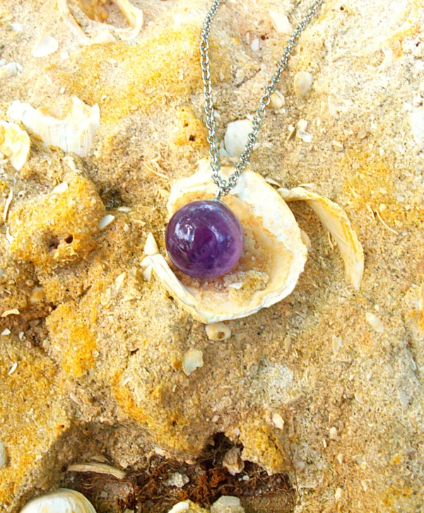 Colgante de cristal de amatista sobre fondo de arena de playa con conchas naturales.