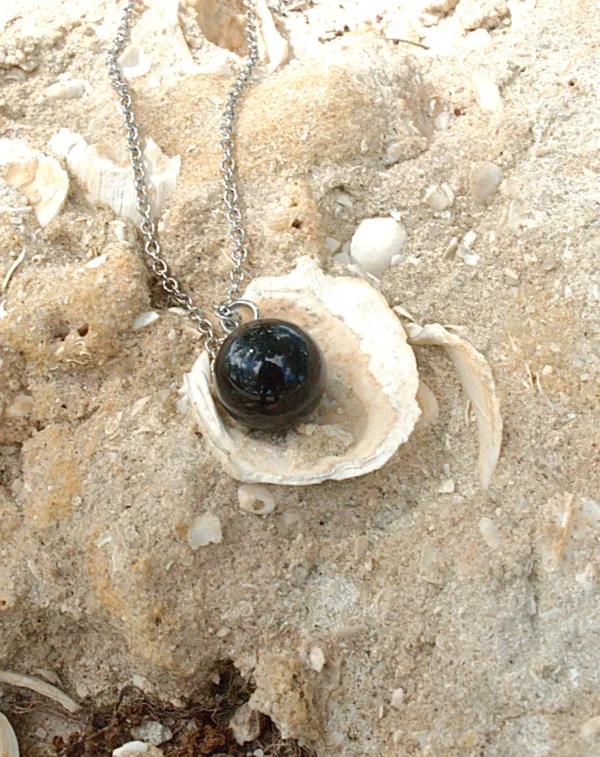 A polished spherical black agate crystal necklace resting in a white seashell on a sandy beach; used as healing crystals for grounding and stress relief.