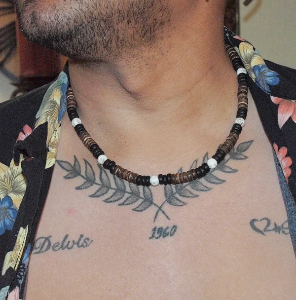 Close-up of a man with a chest tattoo wearing an open black floral tropical shirt and a Queen Conch and coconut shell beaded choker necklace.