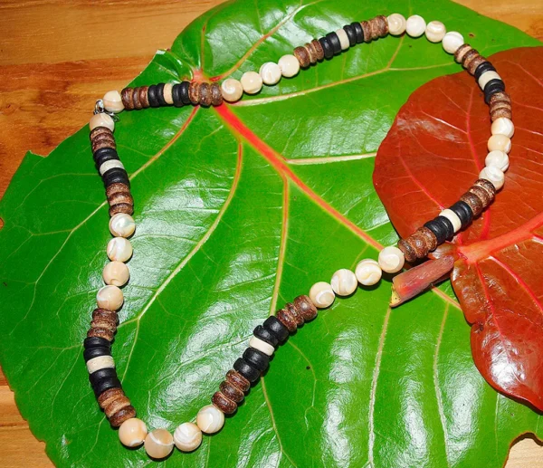 Flat lay product photograph of a handcrafted Trochus shell and coconut shell beaded necklace displayed open on a large green leaf.