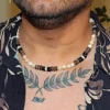 Lifestyle close-up of a man with chest tattoos wearing the Trochus Shell and Coconut Beaded Necklace with a casual black patterned shirt, showcasing summer beach fashion for men.