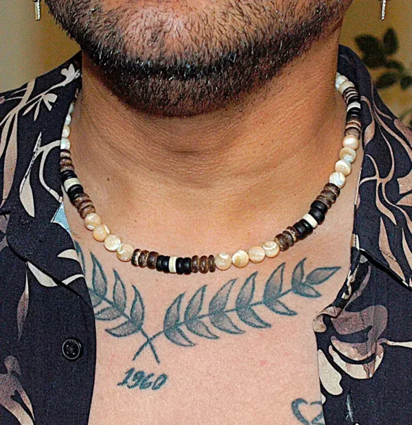Lifestyle close-up of a man with chest tattoos wearing the Trochus Shell and Coconut Beaded Necklace with a casual black patterned shirt, showcasing summer beach fashion for men.