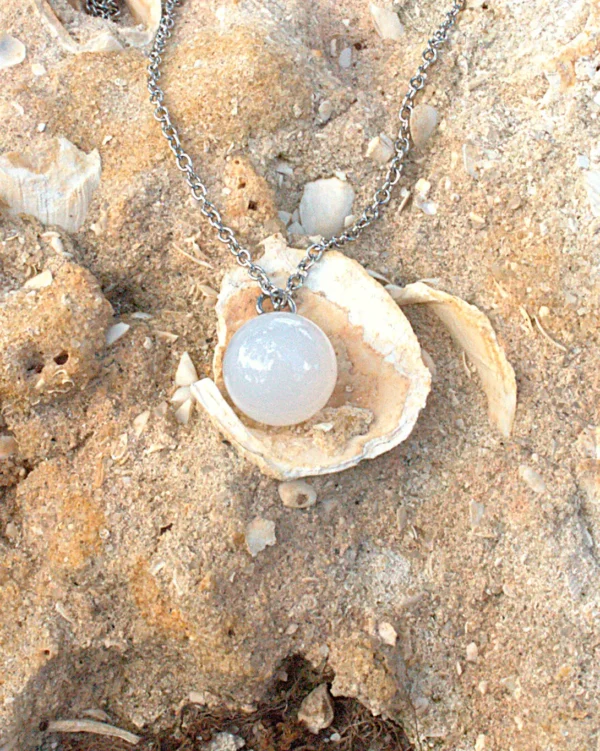 A polished spherical white agate crystal necklace resting on a seashell in the sand; used as healing crystals for anxiety and stress.